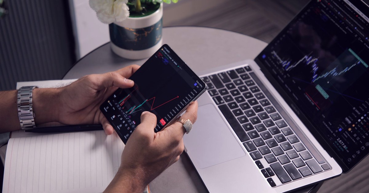 A hand holds a smartphone showing a stock price chart, next to an open laptop displaying candlestick trading charts on a desk with a notebook and a small potted plant