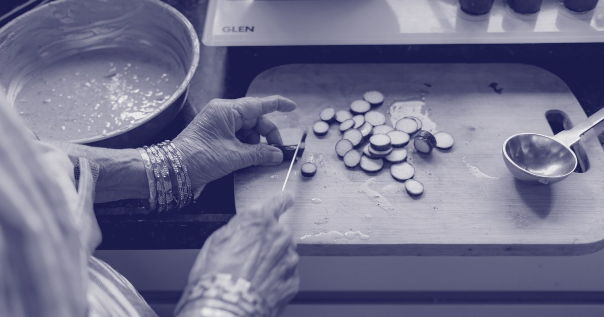 An elderly Indian woman's hands, wearing gold and blue bangles, slice vegetables on a wooden board beside a pot of curry on a home kitchen stove