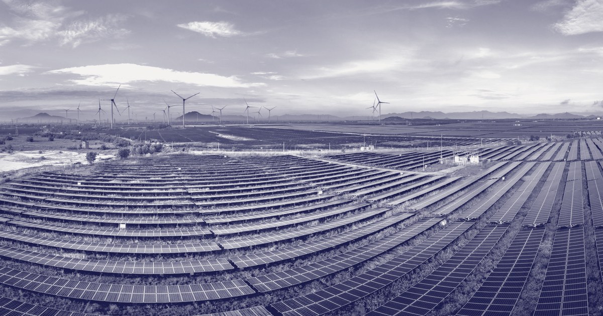 An aerial view of a large solar panel farm with wind turbines on the horizon under a wide sky