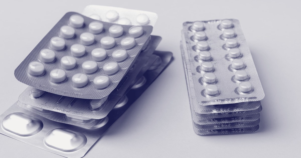 Stacked silver blister packs of tablets and capsules in different colors and shapes, fanned out on a plain background.