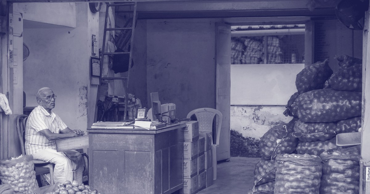 An onion trader seated at his desk in a wholesale shop in India, with large mesh sacks of onions stacked beside cartons of stock