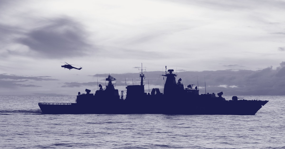 Silhouette of a naval frigate underway at sunset with a helicopter flying overhead