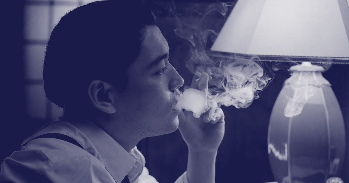A man in a dress shirt and tie exhales a plume of smoke indoors beside a lit table lamp, rendered in navy duotone