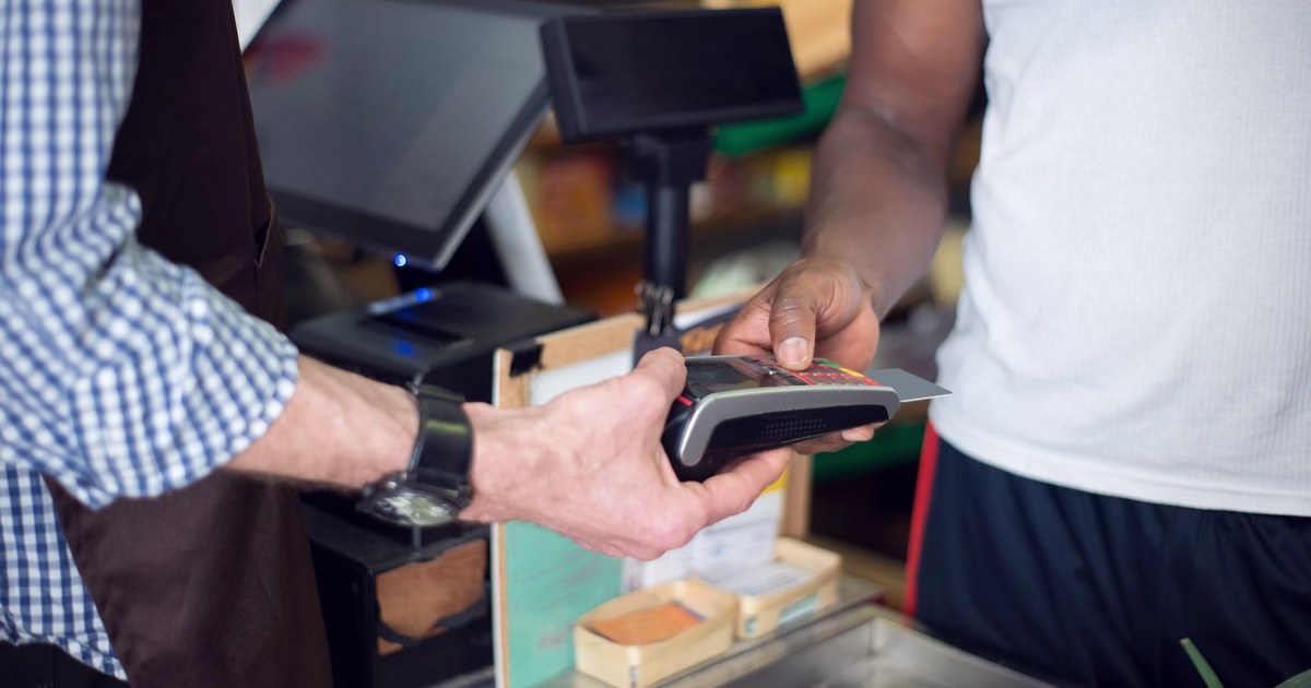 A customer tapping a bank card on a payment terminal at a store counter while a cashier holds the device