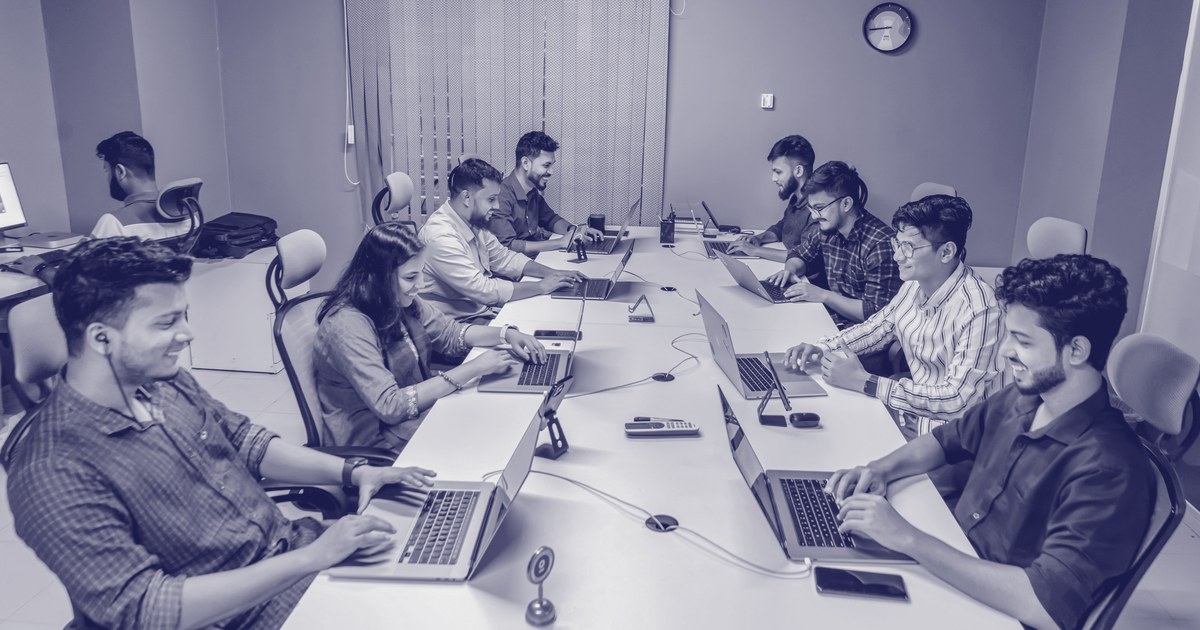 A team of Indian tech and business professionals collaborate on laptops around a long conference table in a modern office