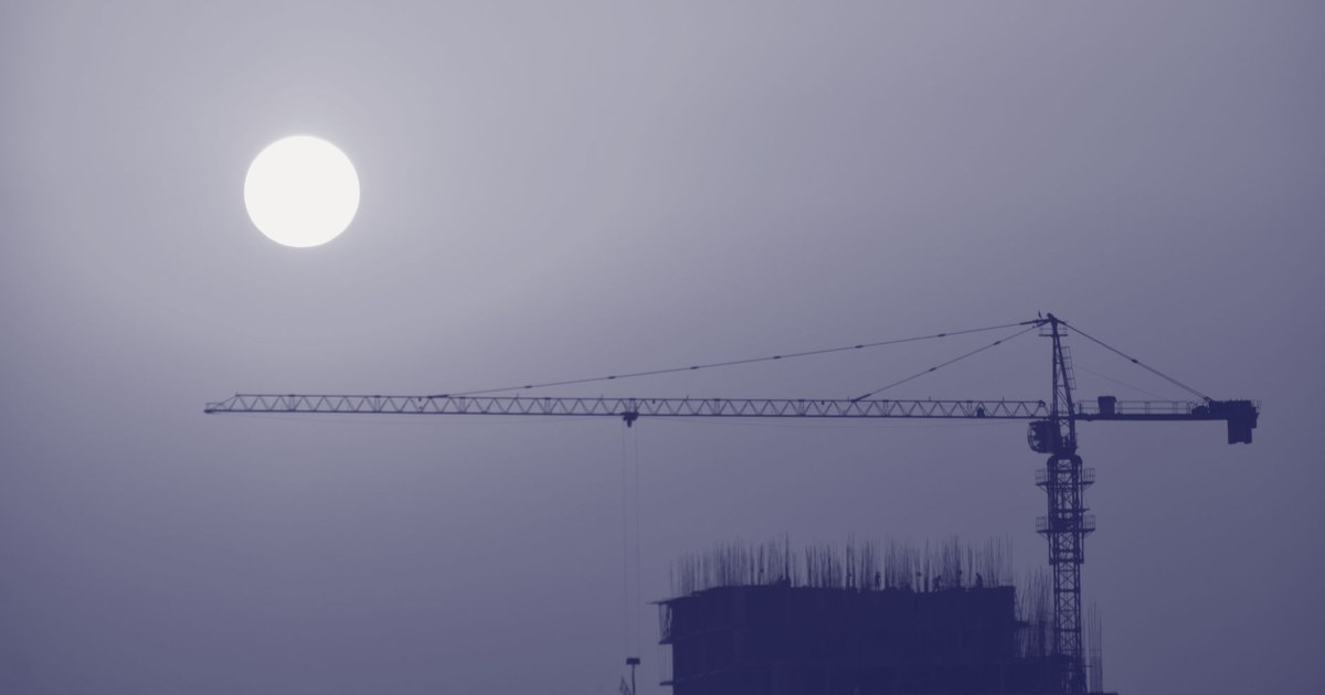 A tower crane and the sun silhouetted in a hazy duotone sky above an unfinished high-rise building in Faridabad, India