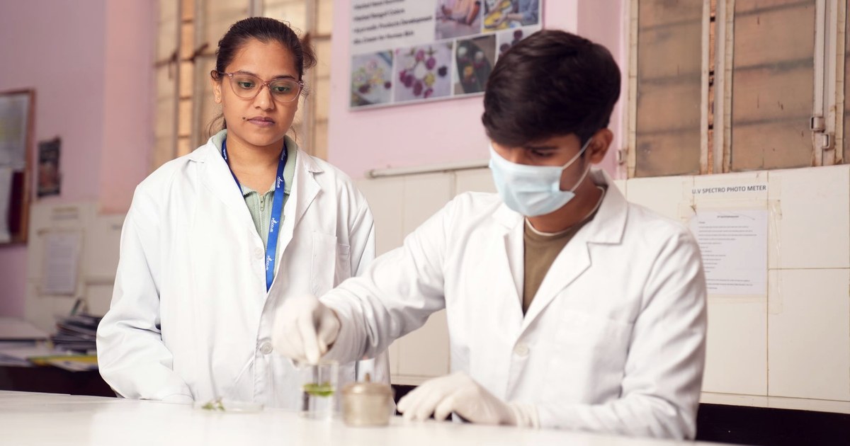 Two young scientists in white lab coats at a laboratory bench in India, one using a dropper to add a green sample to a glass beaker while a colleague looks on