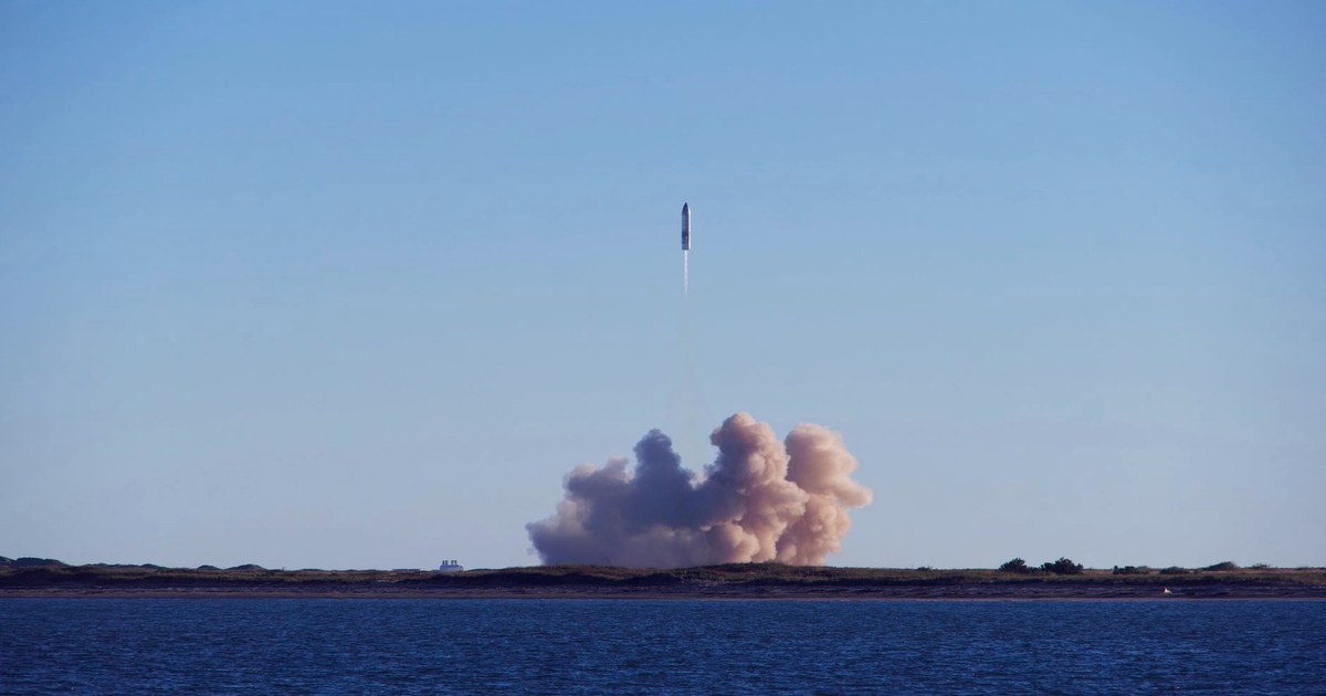 A rocket lifting off over water, trailing a bright exhaust plume into a clear blue sky, with a large plume cloud still billowing at the launch site below