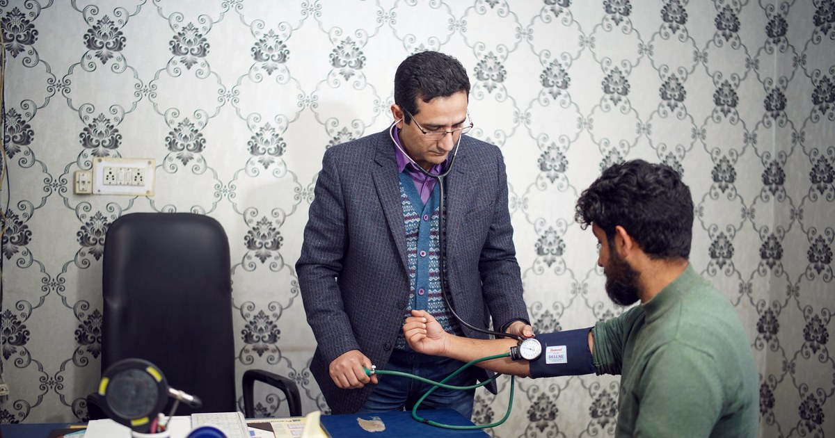 A doctor in an Indian clinic examines a patient with a manual pressure gauge and stethoscope, decorative wallpaper and an Indian-style electrical switchboard visible behind them