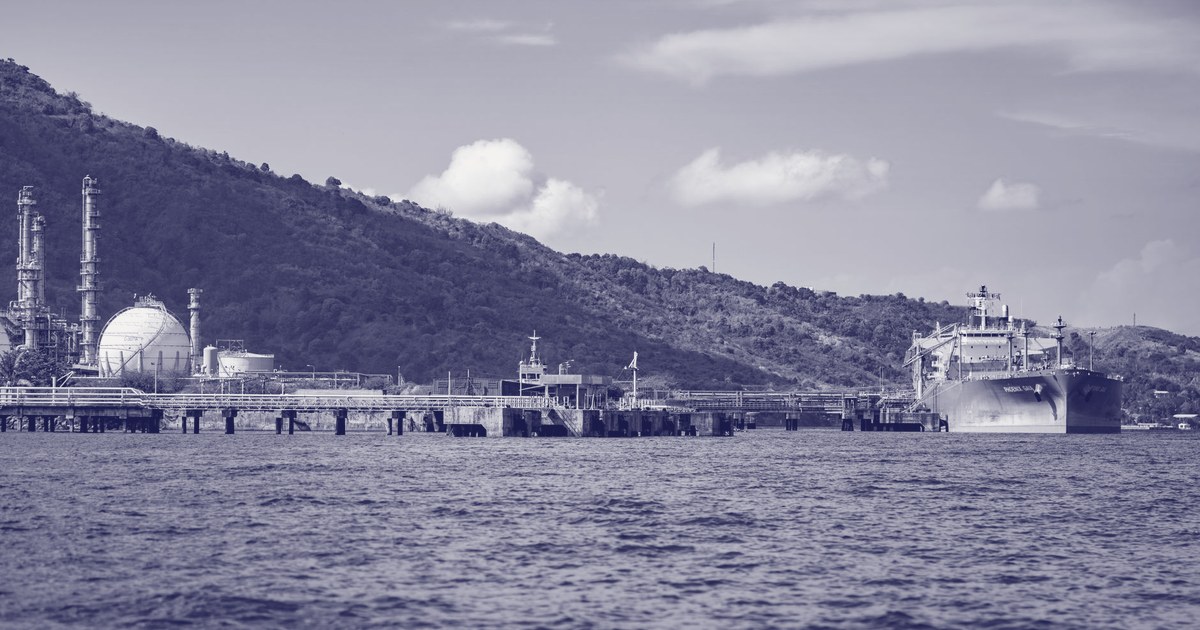 An oil tanker docked at an industrial refinery port, with storage tanks and a hillside in the background