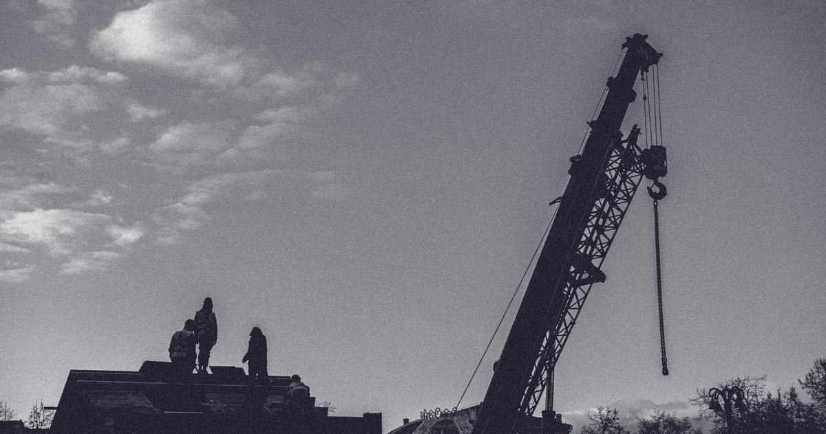 Silhouetted construction workers on a rooftop beside a tall crane against a cloudy sky in Moscow, in black and white