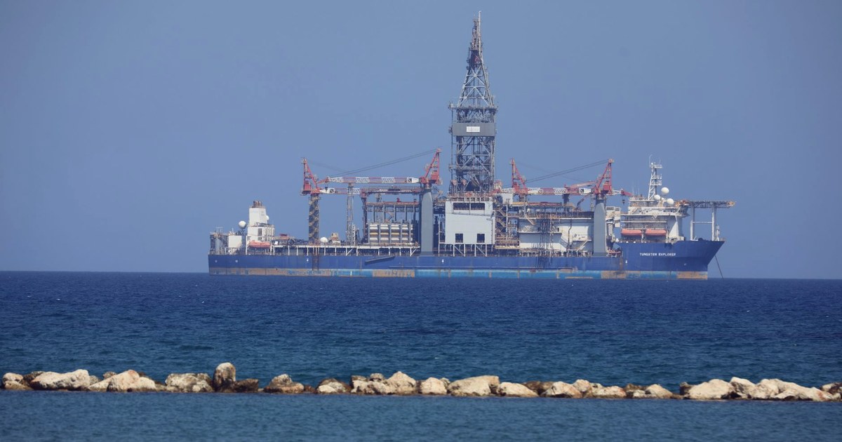 An offshore drillship, the Tungsten Explorer, anchored at sea with its drilling derrick visible, seen from a rocky shoreline