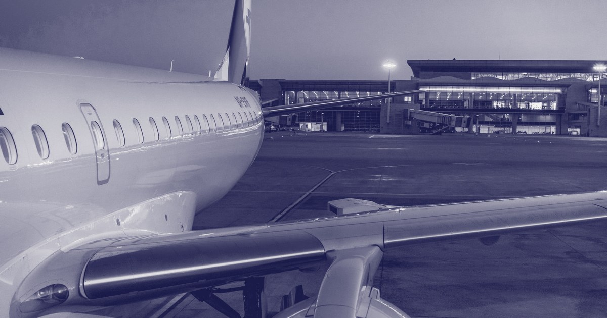 Close-up of a commercial airplane's fuselage and wing at an airport gate at dusk, with the terminal building lit up in the background