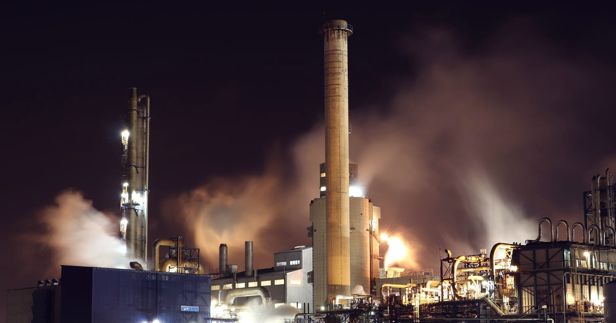 A heavy-industry plant at night, with a tall chimney and steam clouds lit by floodlights
