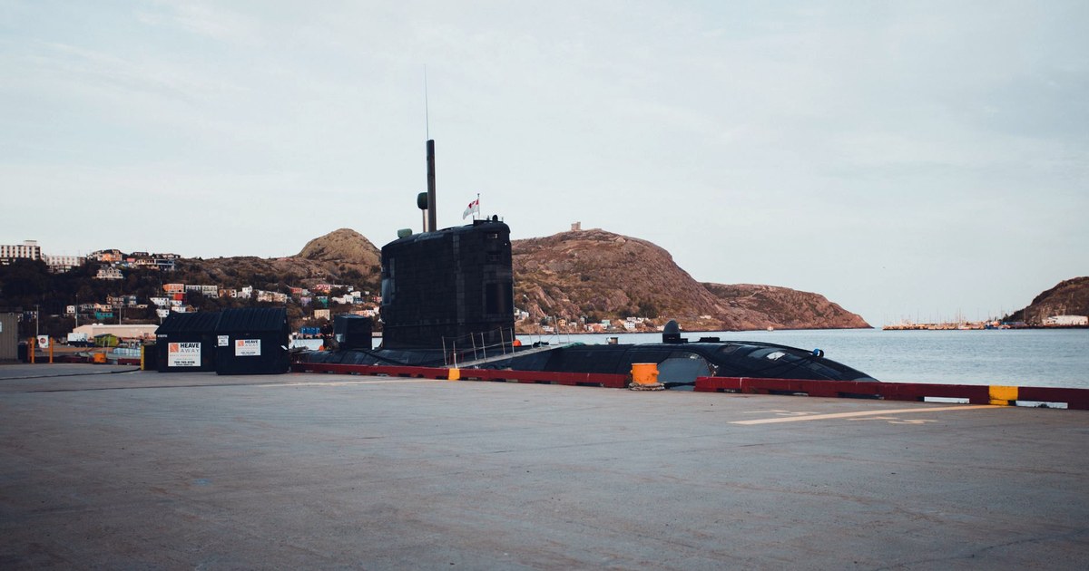 A large black conventional submarine docked at a harbor pier, with hills and a coastal town in the background
