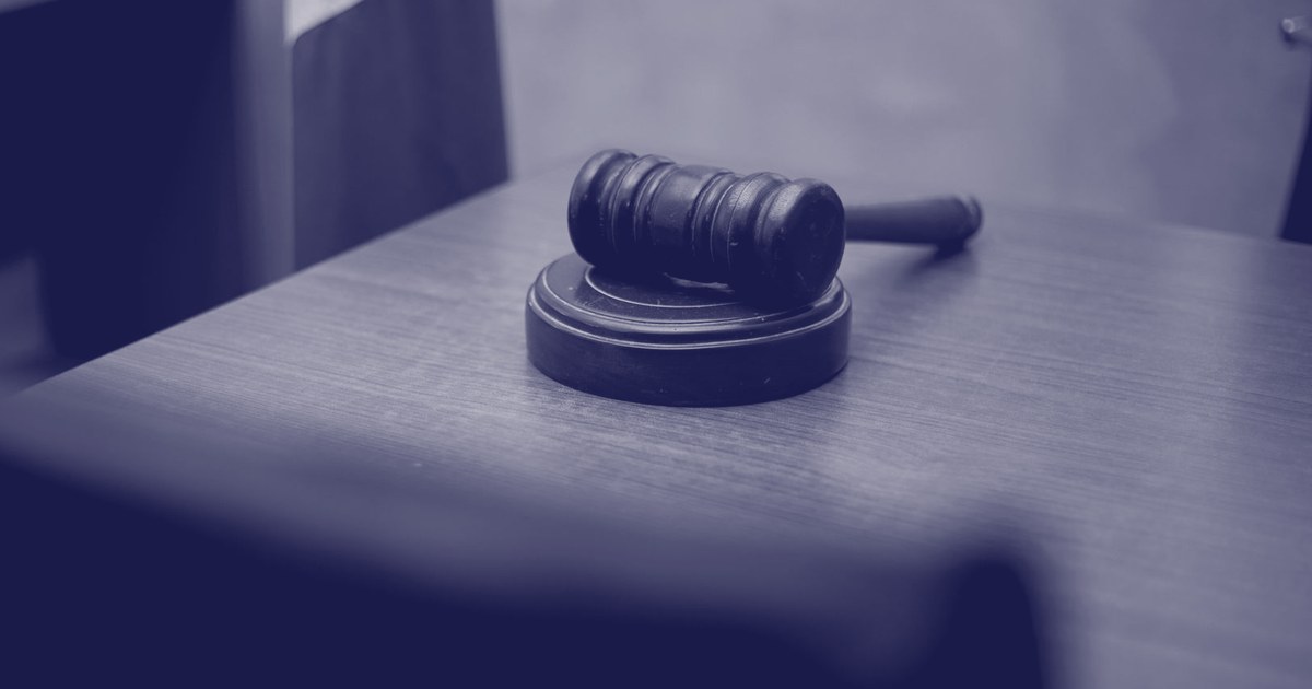 A judicial gavel resting on a wooden courtroom desk, with a flag visible blurred in the background