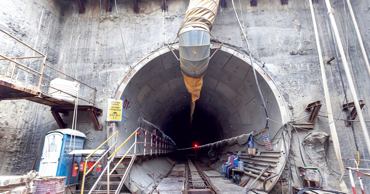 The concrete entrance of an under-construction railway tunnel in India, with a ventilation duct, safety signage in Hindi and English, and workers seated beside the rail track leading into the dark bore
