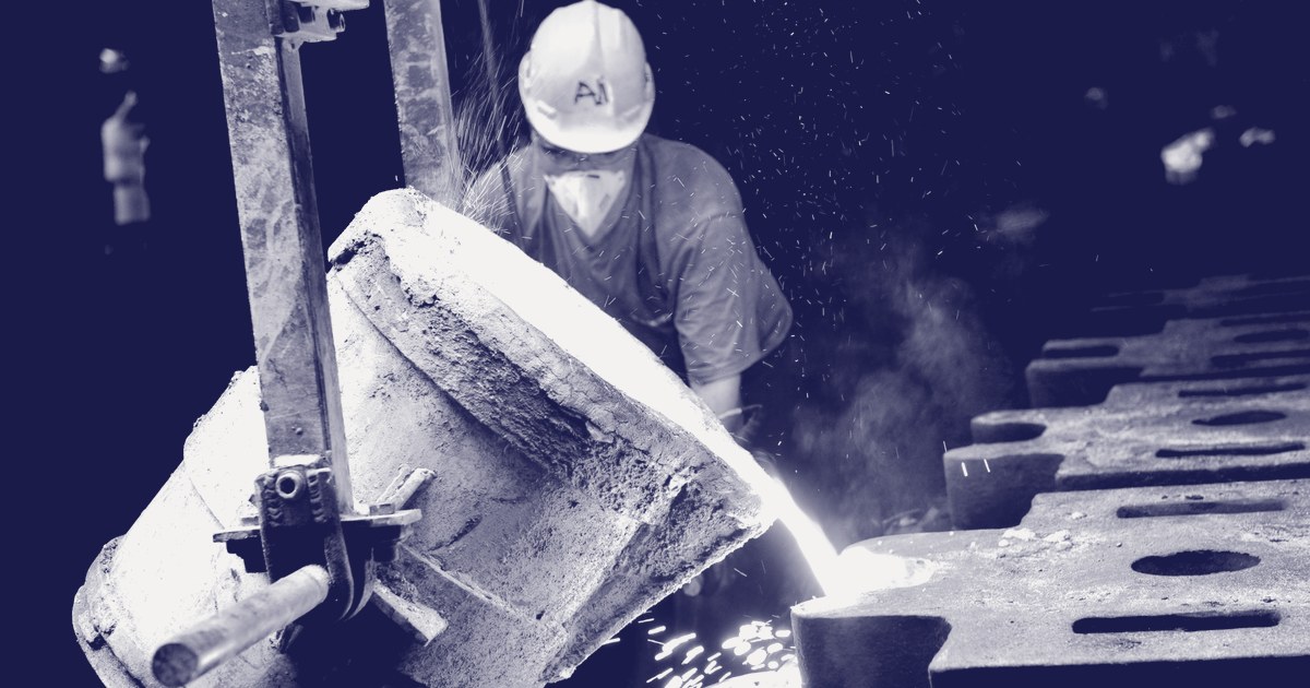 A foundry worker in a hard hat and respirator pours molten metal from a crucible into a casting mold, sparks flying