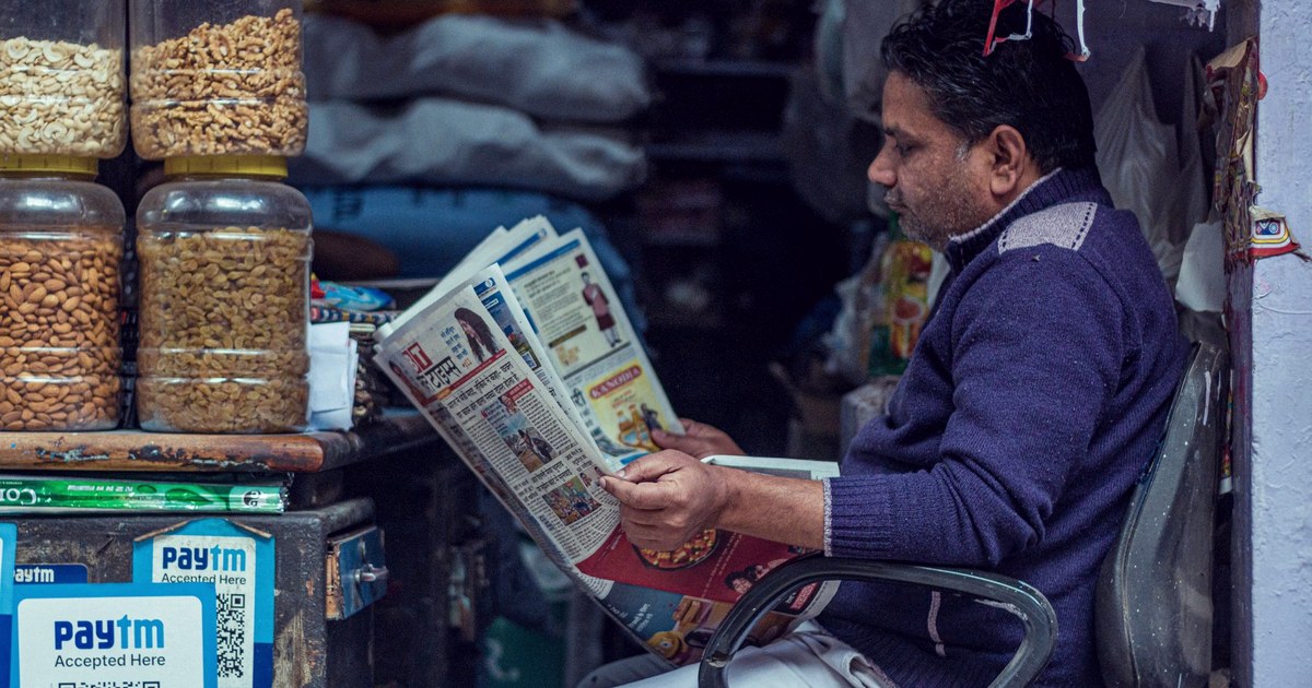 A shopkeeper at a dry fruit and grocery stall reads a newspaper beside a Paytm UPI QR code payment sign