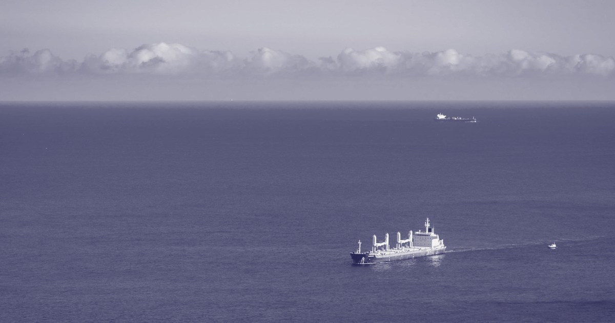 A cargo ship crossing open ocean, seen from above, with a smaller vessel in the distance near the horizon