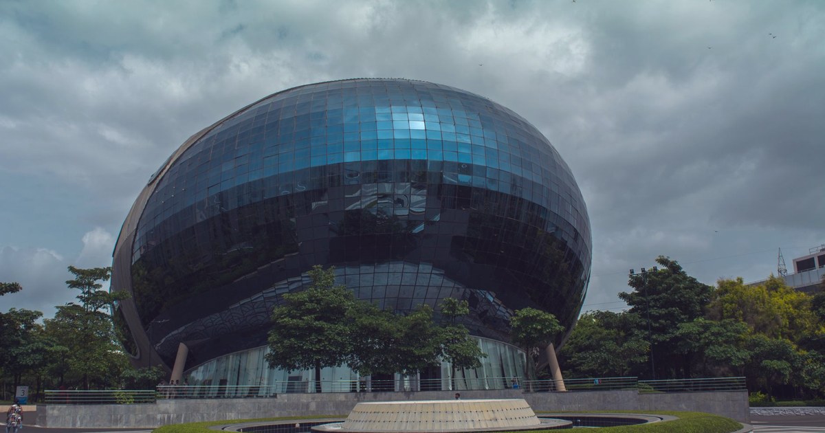 A distinctive egg-shaped glass office building with a mirrored, reflective facade, surrounded by trees at a technology park in India