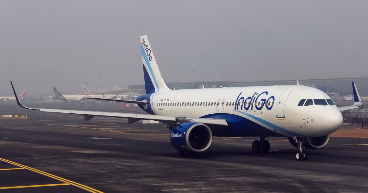 An IndiGo passenger Airbus aircraft taxiing on the tarmac at a foggy airport, with other aircraft visible in the background