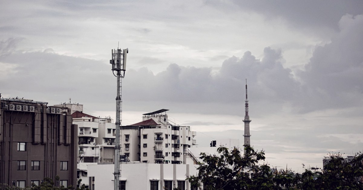A mobile network tower rising above apartment buildings in an Indian city under a cloudy sky, with a taller telecom mast in the background