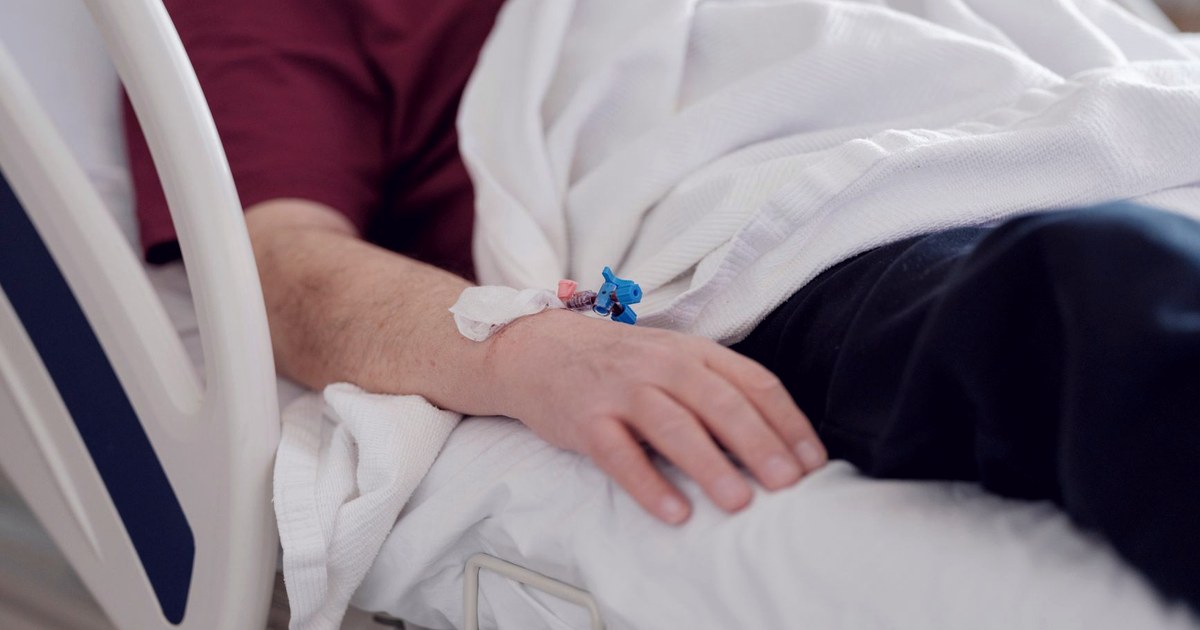 A hospital patient's arm resting on a pillow with an IV cannula taped in place, in a hospital bed
