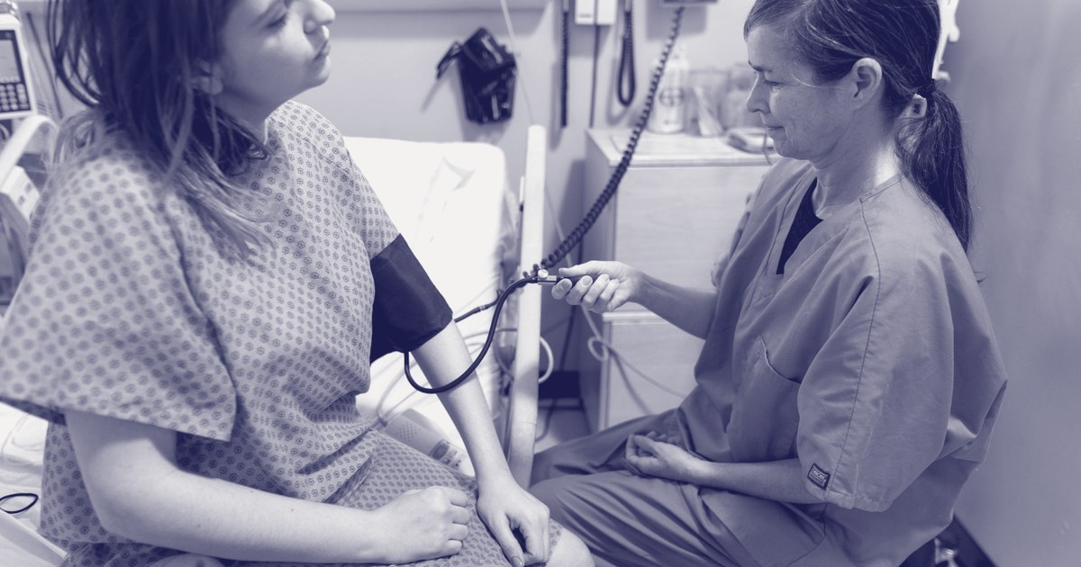 A nurse in scrubs checks a seated patient's vitals with an arm cuff in a hospital room