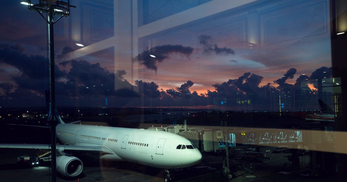 A white passenger jet connected to an airport terminal by an airbridge at dusk, seen through glass under a purple and orange sky
