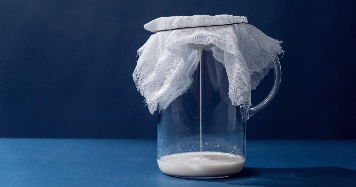 A glass jar covered with cheesecloth catching a thin stream of milk being strained, against a dark blue background