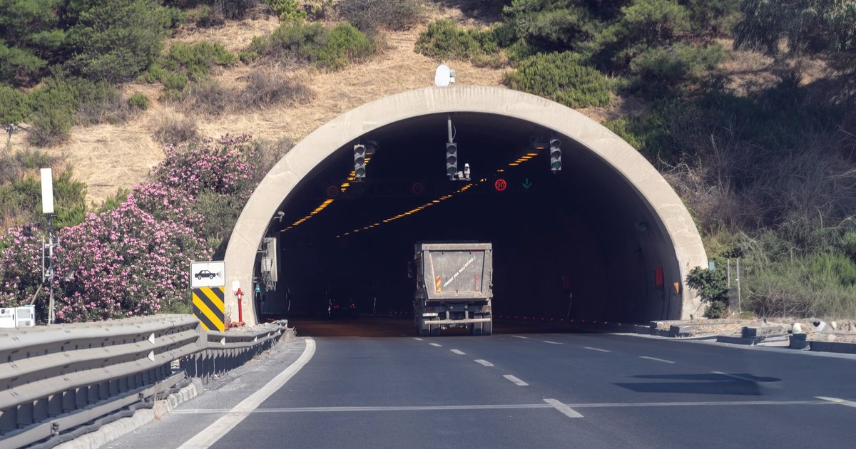 A highway tunnel entrance cut into a green hillside, with a truck approaching the dark tunnel mouth on a two-lane carriageway