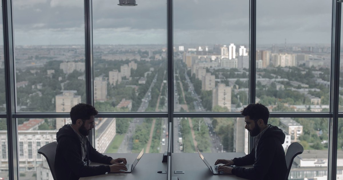 Two office workers at laptops in a glass-walled office with a wide view of a city below