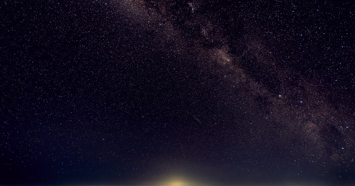 The Milky Way arcing across a dark sky, fading into a warm dome of artificial light glowing up from a city on the horizon