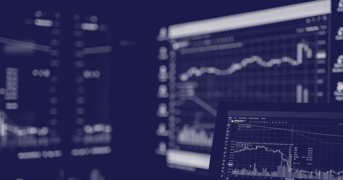 Multiple computer and laptop screens displaying stock market candlestick charts and trading data in a dark room