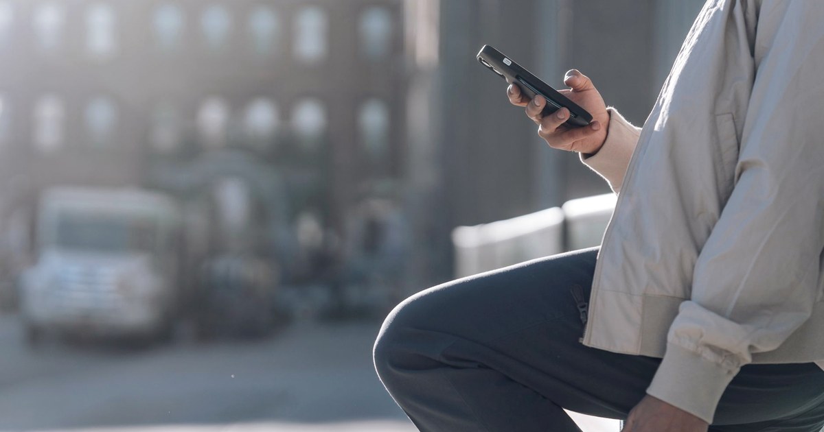 A hand holding a smartphone with its screen turned away from view, standing on a city street in daylight