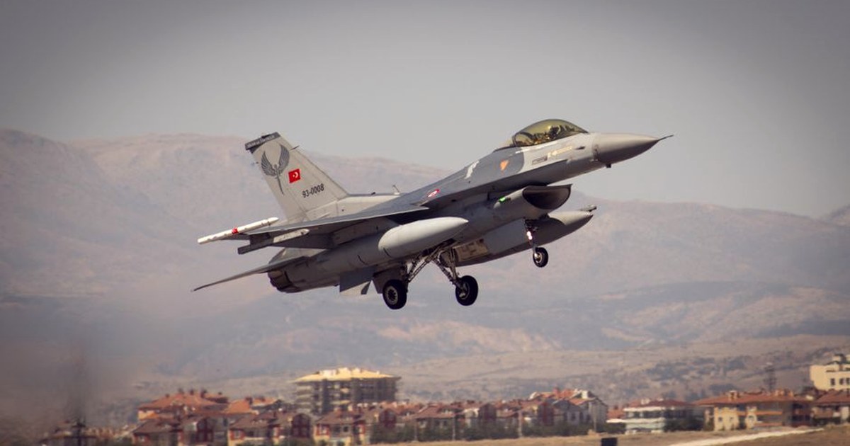 A Turkish Air Force F-16 fighter jet taking off, with a residential town and mountains in the background