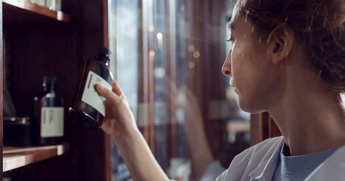 A pharmacist in a white coat examining an amber medicine bottle on a wooden shelf, backlit by a window