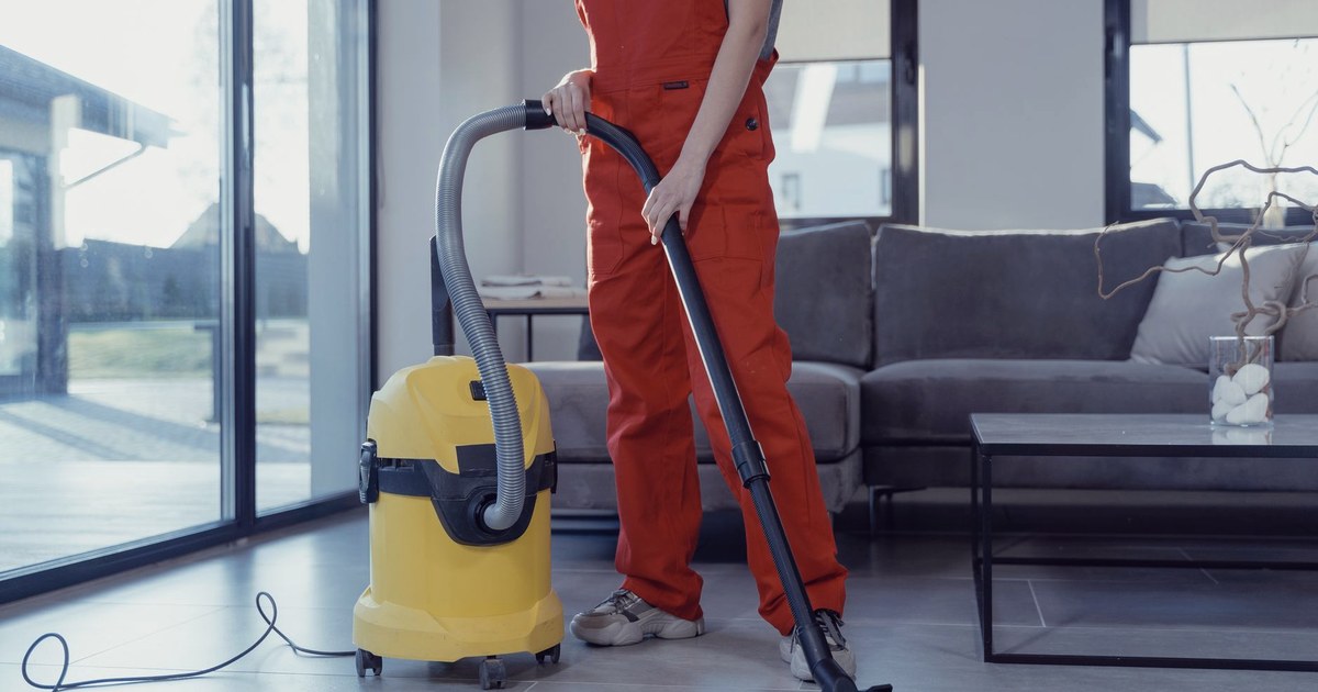 A professional cleaner in red coveralls vacuuming a modern living room with a yellow canister vacuum cleaner