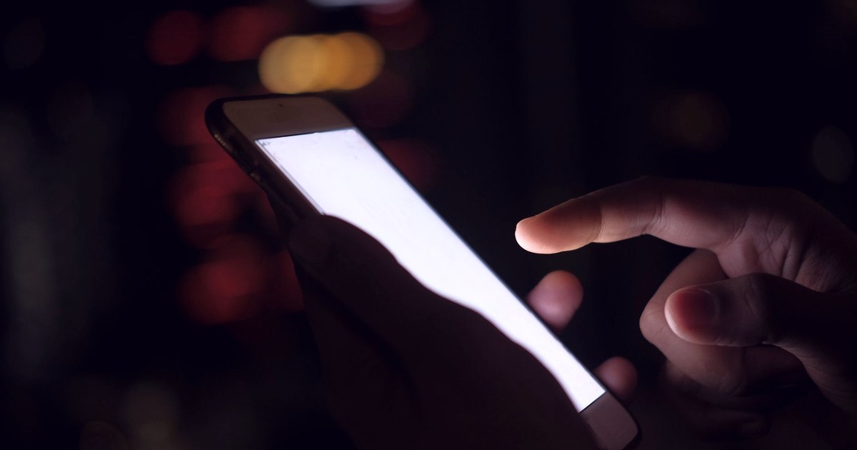 A hand touches the glowing screen of a smartphone in the dark, with blurred red and amber city lights in the background