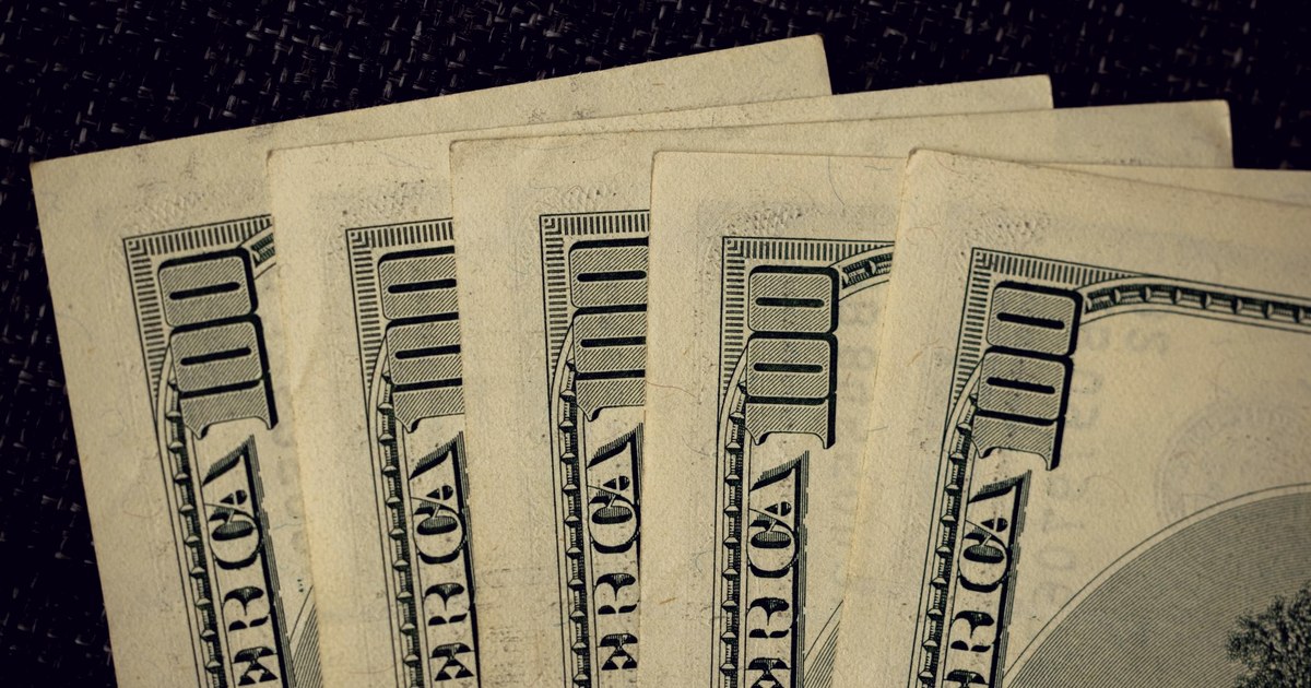 A fanned stack of US one-hundred dollar bills photographed close up against a dark woven surface