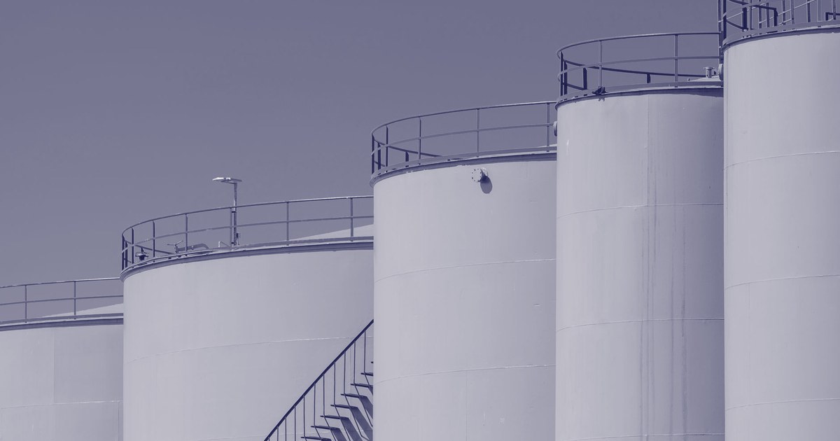 A row of large industrial fuel storage tanks with railings and a steel staircase, in a muted violet duotone