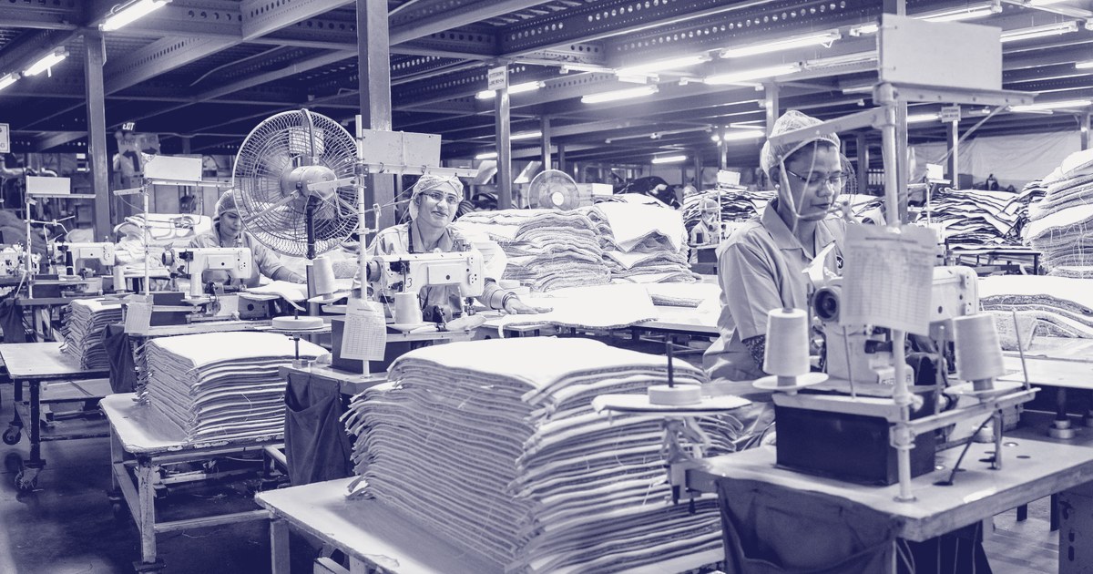Workers at sewing machines beside tall stacks of fabric on a large factory floor, in a violet duotone treatment