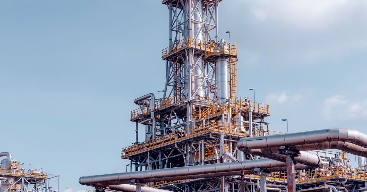A tall industrial gasification and processing tower with layered pipelines and yellow platform railings against a clear sky