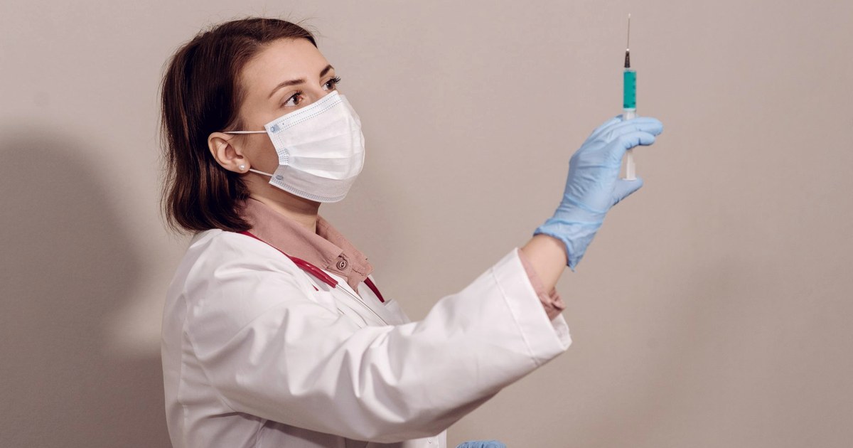 A masked doctor in a white coat and blue gloves holds a syringe up to the light against a beige wall