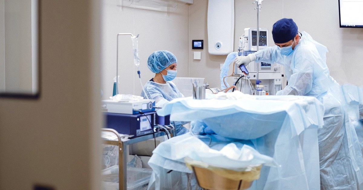 A surgical team in blue gowns and caps working around a draped operating table under equipment monitors in a sterile operating room