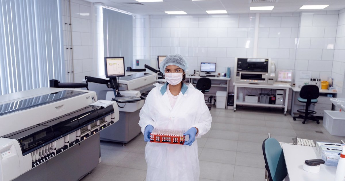 A laboratory technician in a mask and hairnet holds a rack of DNA sample vials in a clinical genetics laboratory