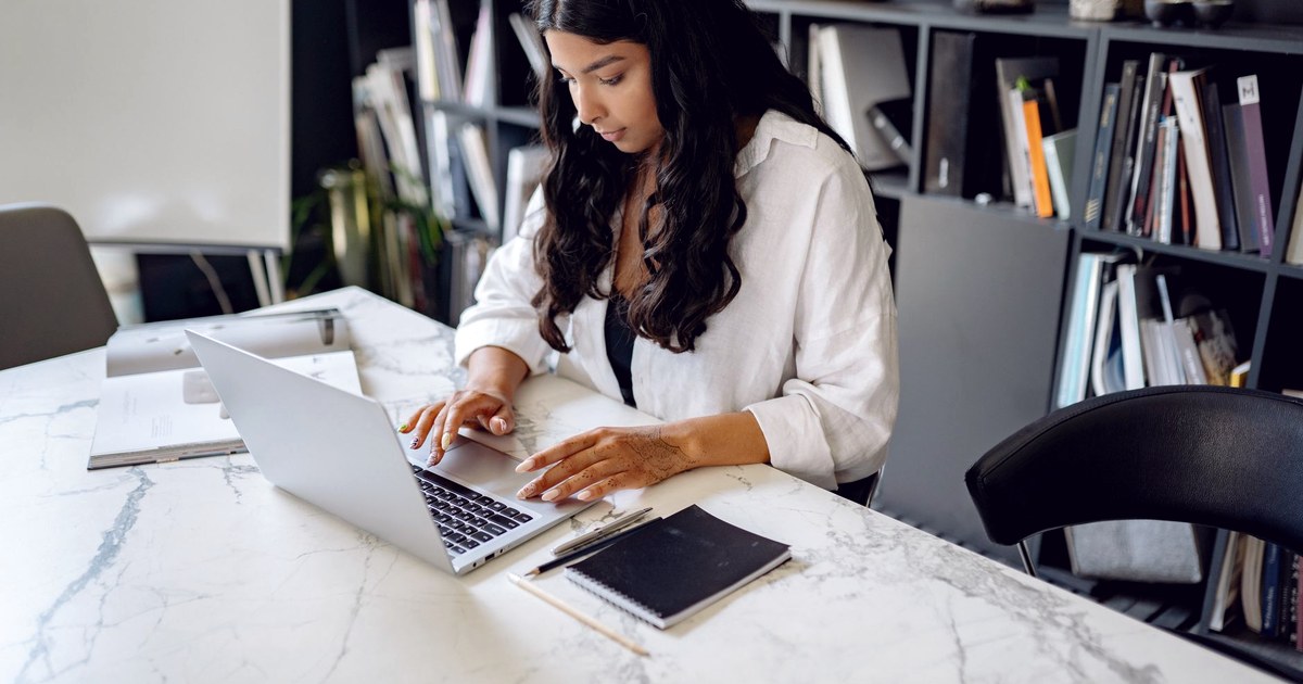A woman working on a laptop at a marble table in an office, with a notebook and bookshelves behind her