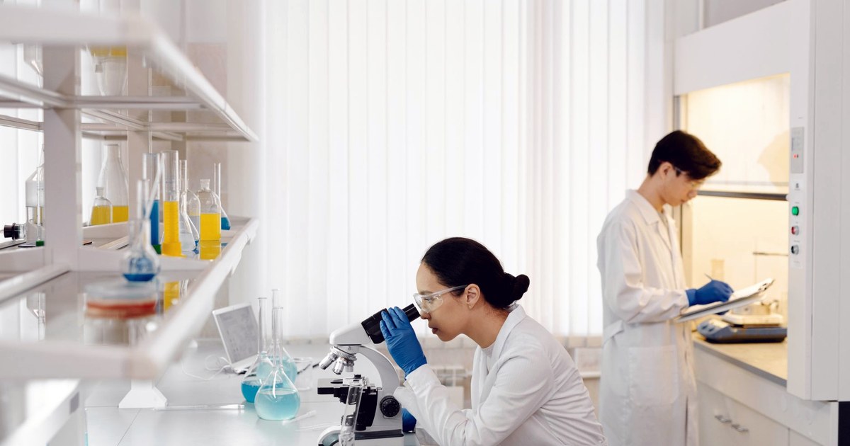 Two researchers in white coats working in a bright laboratory, one looking through a microscope while the other writes on a clipboard