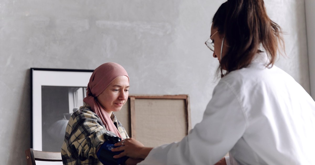 A doctor in a white coat checks a seated woman's arm-cuff hypertension reading, with the woman wearing a headscarf, in a clinic setting